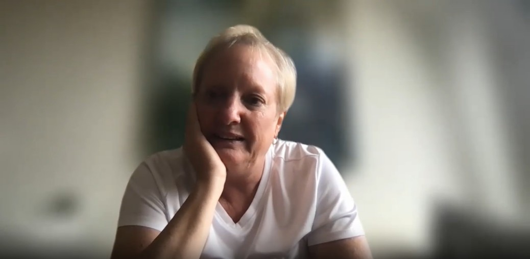 A picture of a blonde woman with short hair with her hand on her chin looking contemplative. Her background is blurred using Zoom.